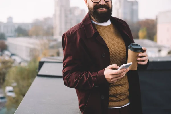 Vista cortada de homem adulto elegante em óculos segurando café para ir e usando smartphone no telhado — Fotografia de Stock