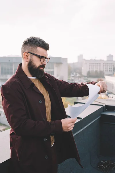 Ernsthafte erwachsene männliche Architekt in Brille mit Blaupause und Arbeit an einem Projekt auf dem Dach — Stockfoto
