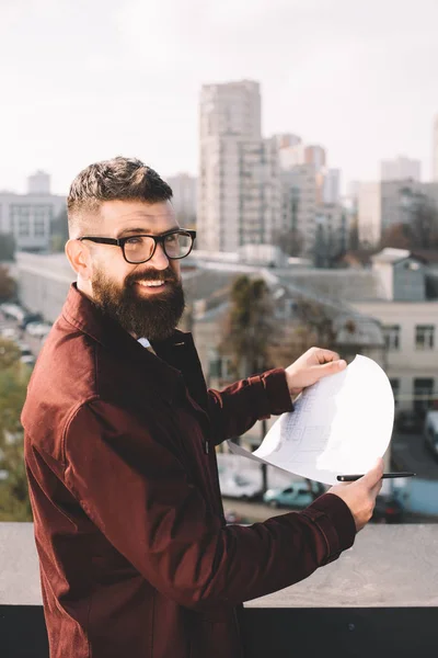Sorridente architetto maschio adulto in occhiali che tiene cianografia sul tetto con bella vista — Foto stock