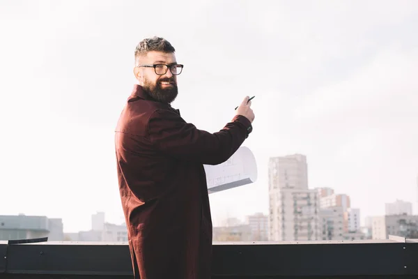 Architecte masculin adulte souriant dans des lunettes tenant le plan directeur sur le toit — Photo de stock