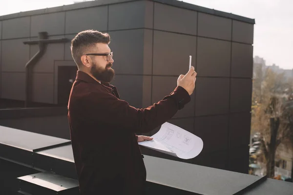 Serious adult male architect in glasses taking photo and holding blueprint on rooftop — Stock Photo
