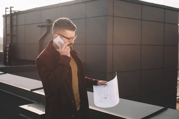 Serious adult male architect in glasses talking on smartphone and holding blueprint on rooftop — Stock Photo