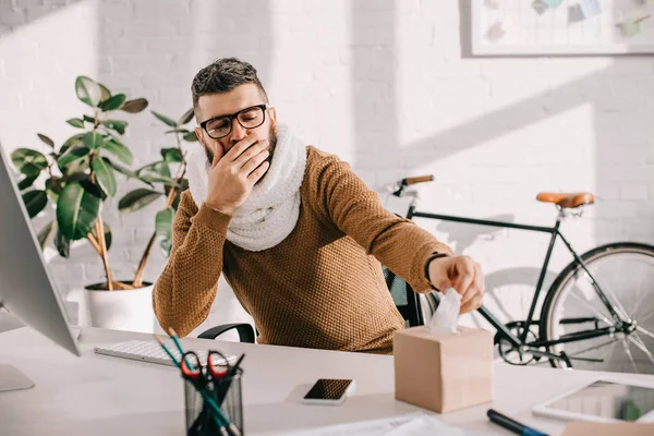 Empresário doente em cachecol de malha sentado na mesa de escritório, tosse e estendendo a mão para tecidos — Fotografia de Stock