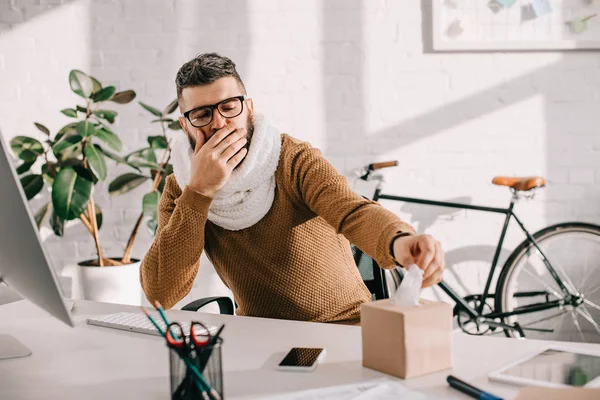 Hombre de negocios enfermo en bufanda de punto sentado en el escritorio de la oficina, tosiendo y buscando pañuelos - foto de stock