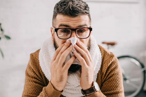 Portrait of sick man in knitted scarf sitting in office, sneezing and covering mouth with tissue — Stock Photo
