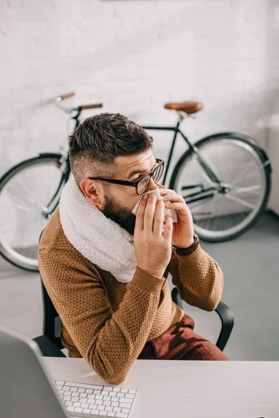 Больной бизнесмен в трикотажном шарфе сидит в офисе, чихает и покрывает рот тканью — стоковое фото