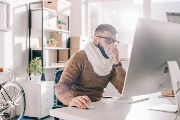 Homme d'affaires malade en écharpe tricotée assis dans le bureau, éternuant et couvrant la bouche avec du tissu — Photo de stock