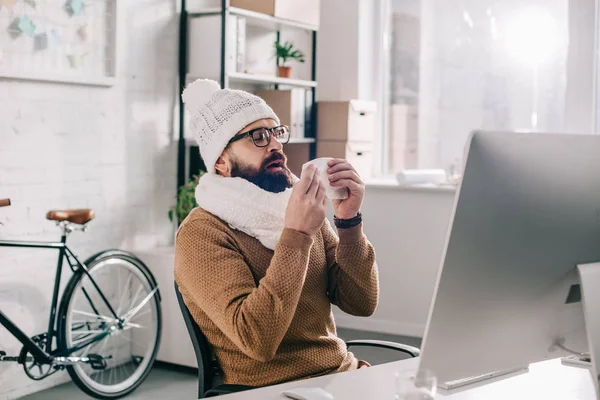 Sick businessman in scarf and knitted hat sitting at office desk and sneezing — Stock Photo