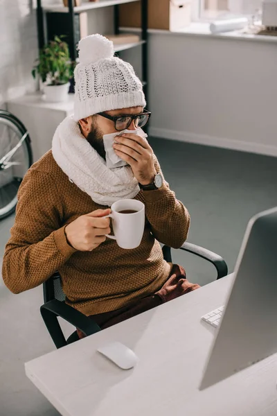 Sick businessman in scarf and knitted hat sitting at office desk, holding cup of tea and sneezing — Stock Photo