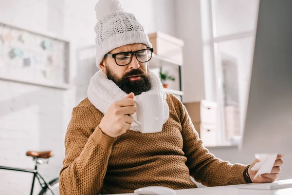 Uomo d'affari malato in sciarpa e cappello a maglia che tiene tazza di tè e tessuto sul posto di lavoro — Foto stock