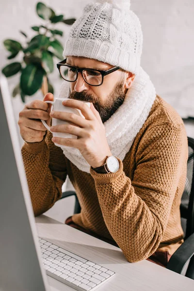 Sick businessman in scarf and knitted hat working at computer desk and drinking tea in office — Stock Photo