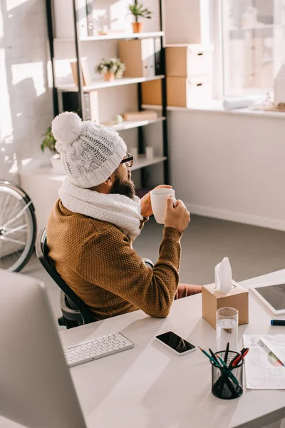 Sick businessman in scarf and knitted hat sitting at office desk and holding cup of tea — Stock Photo