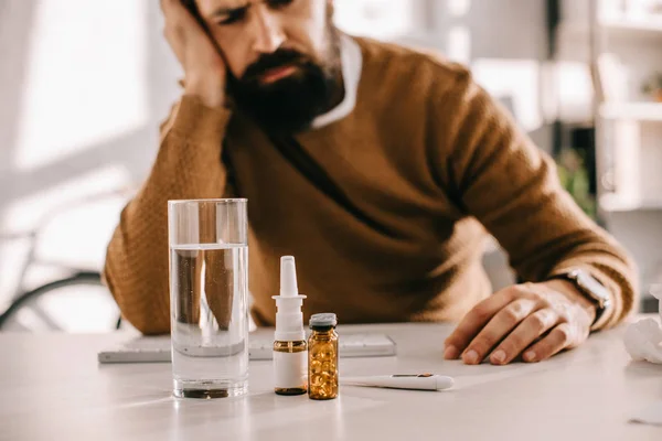 Selektiver Fokus eines kranken Geschäftsmannes, der mit Medikamenten, Nasenspray und einem Glas Wasser am Schreibtisch sitzt — Stockfoto