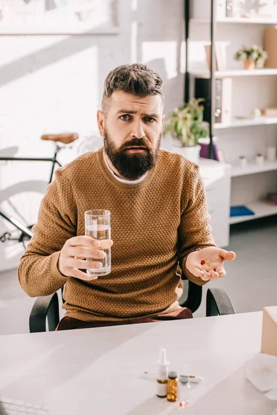 Больной бизнесмен сидит за рабочим столом, смотрит в камеру, держа в руках стакан воды и лекарства — стоковое фото
