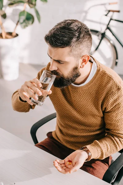 Angle view of sick businessman sitting at office desk, holding medicine and drinking water at workplace — Stock Photo