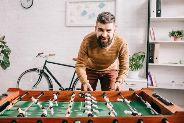 Lächelnder schöner erwachsener Mann, der in die Kamera schaut und zu Hause Tischkicker spielt — Stockfoto