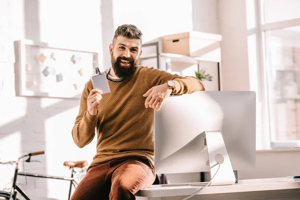 Homem de negócios adulto barbudo alegre sentado na mesa e apontando para etiqueta de segurança em branco no escritório — Fotografia de Stock