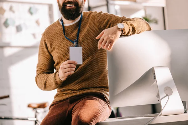 Обрізаний вид бородатого дорослого бізнесмена, що сидить на столі і тримає порожню мітку безпеки в офісі — стокове фото
