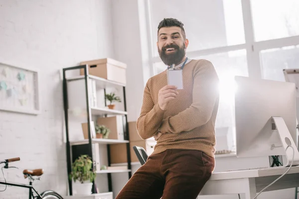 Smiling bearded adult businessman sitting on office desk and holding blank security tag at workplace — Stock Photo