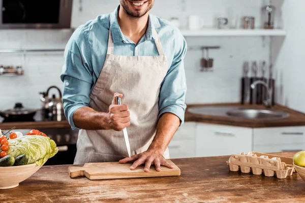 Tiro cortado de jovem sorridente em avental segurando faca e mão em tábua de corte na cozinha — Fotografia de Stock