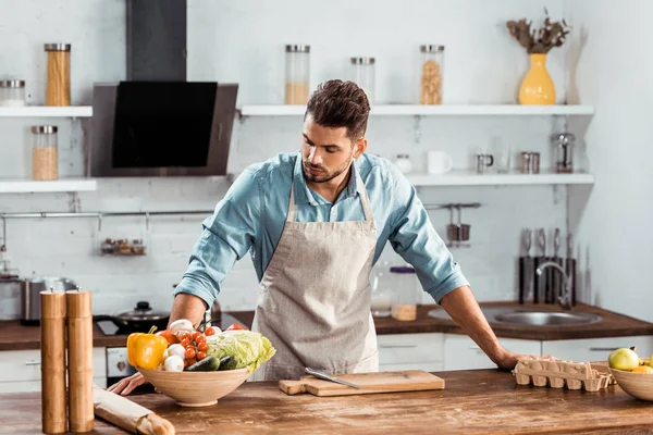 Handsome young man in apron leaning at table and looking at fresh vegetables in kitchen — Stock Photo