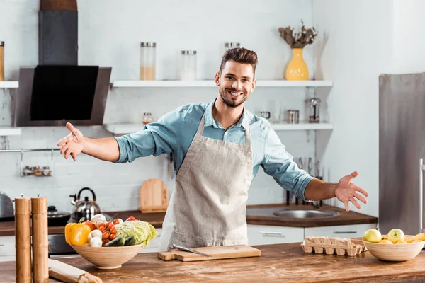 Joyeux jeune homme dans le tablier avec les bras ouverts souriant à la caméra dans la cuisine — Photo de stock