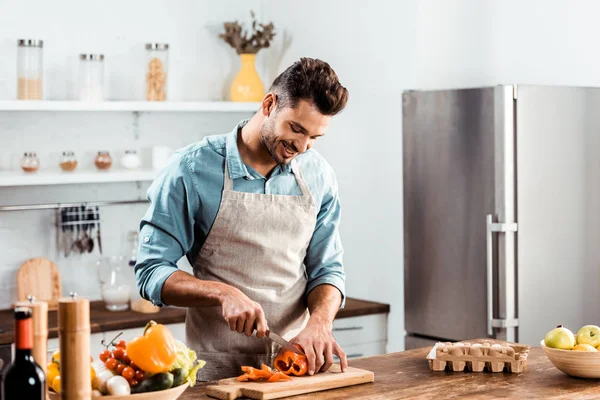 Улыбающийся молодой человек в фартуке режет свежий перец на кухне — стоковое фото