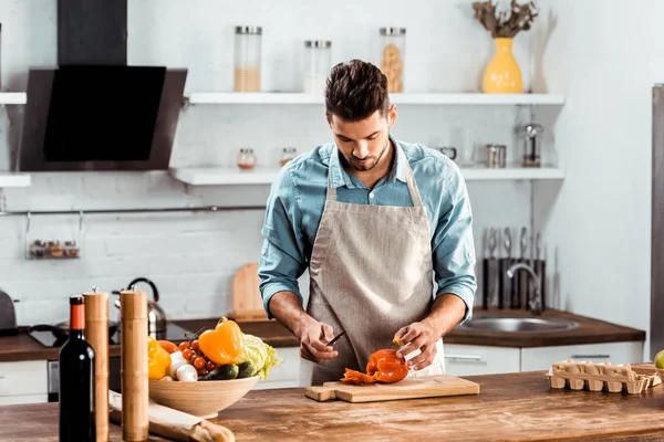 Young man in apron cutting fresh pepper in kitchen — Stock Photo