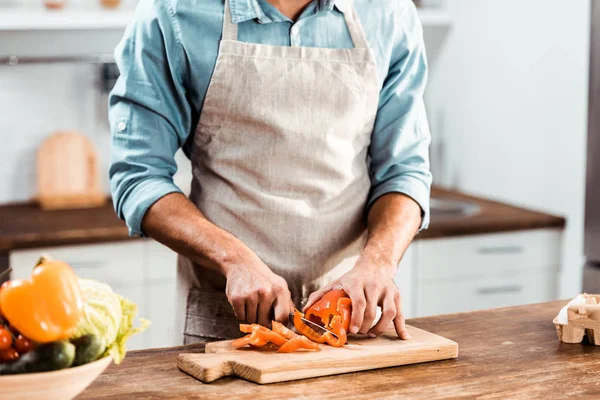 Sección media del joven en delantal corte de pimienta fresca en la cocina - foto de stock