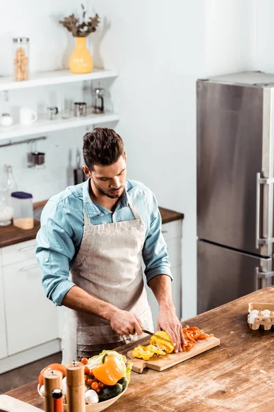 High angle view of young man in apron cutting fresh peppers in kitchen — Stock Photo