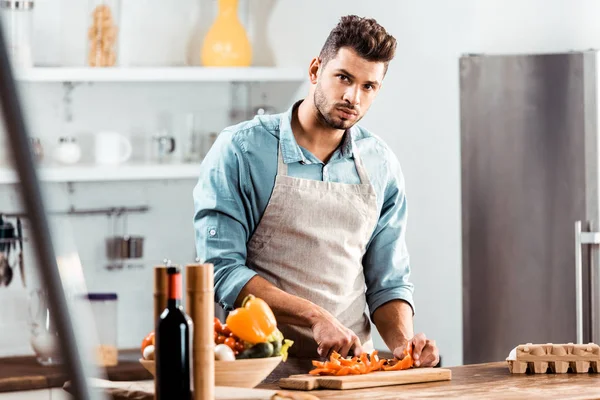 Handsome young man in apron cutting vegetables and looking at camera in kitchen — Stock Photo