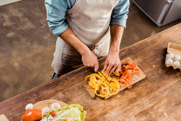 Cropped shot of man in apron cutting fresh peppers in kitchen — Stock Photo