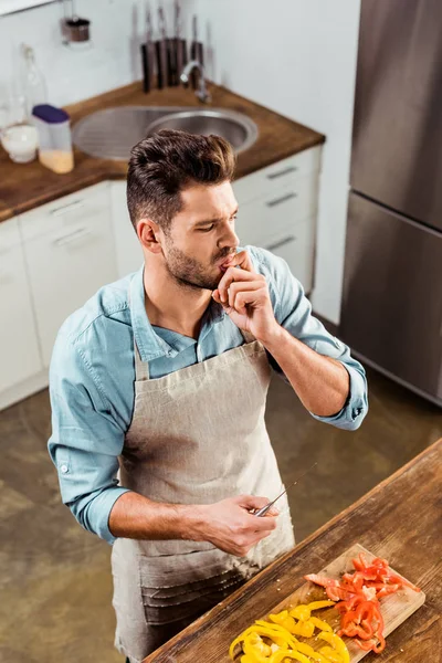 Vista ad alto angolo dell'uomo in grembiule con coltello e dito leccare mentre taglia le verdure — Foto stock