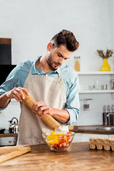 Young man in apron adding pepper with mill into vegetable salad — Stock Photo
