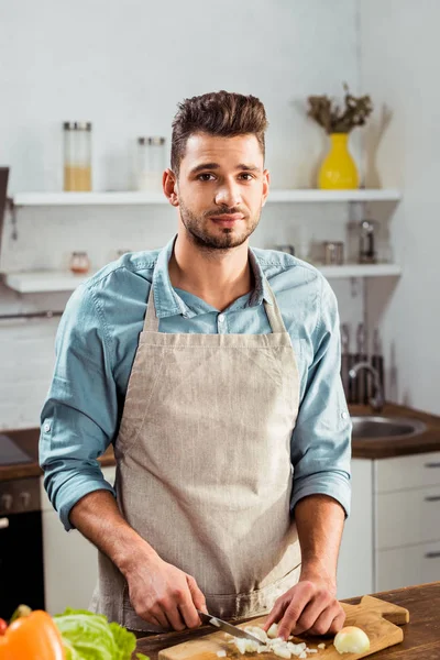 Schöner junger Mann in Schürze, der Zwiebel hackt und in der Küche in die Kamera schaut — Stockfoto