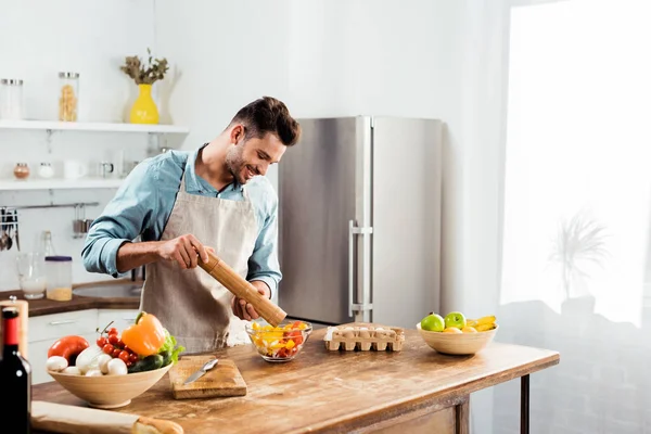 Schöner lächelnder junger Mann, der Pfeffer mit Mühle in den Salat gibt — Stockfoto