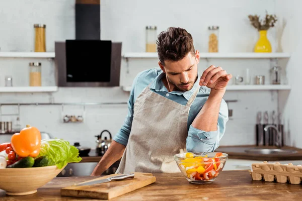 Schöner junger Mann in Schürze, der Gemüsesalat salzt, während er in der Küche kocht — Stockfoto