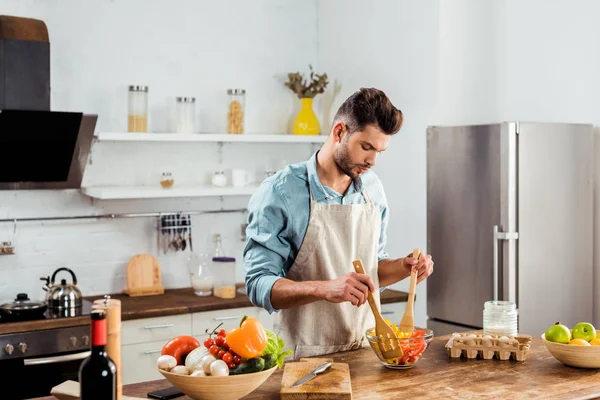 Handsome young man in apron cooking vegetable salad in kitchen — Stock Photo