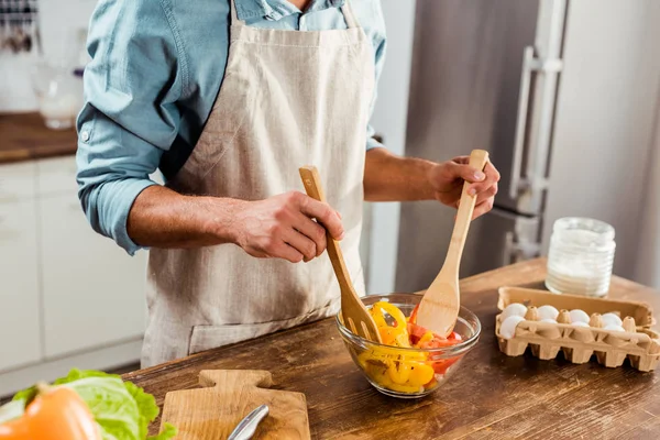 Tiro cortado de jovem em avental misturando salada de legumes na cozinha — Fotografia de Stock