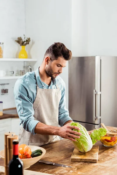 Молодий чоловік в фартусі тримає капусту і готує на кухні — стокове фото