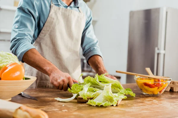 Tiro recortado de hombre joven en delantal de corte de verduras en la cocina - foto de stock