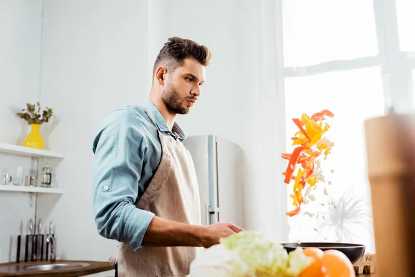 Vista lateral del joven en delantal sacudiendo sartén con verduras - foto de stock