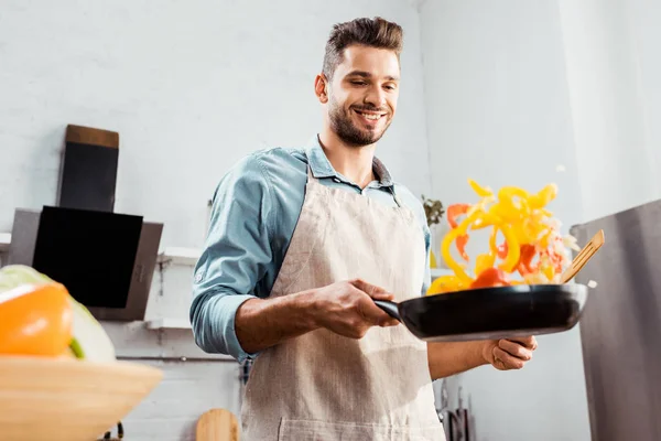 Vista de ángulo bajo del joven sonriente en delantal sosteniendo sartén con verduras — Stock Photo