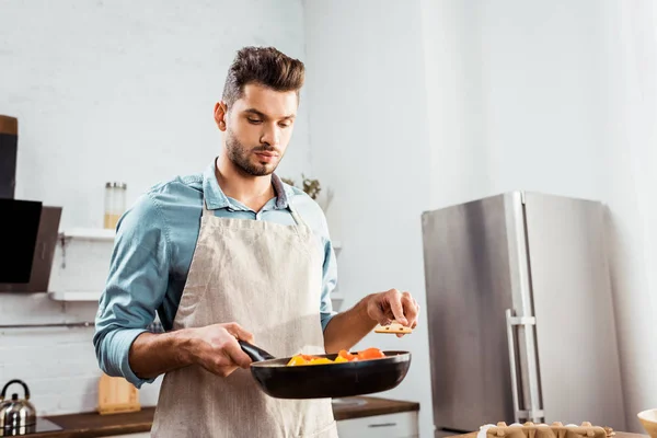 Handsome young man in apron holding frying pan with vegetables — Stock Photo