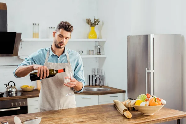 Bel giovanotto in grembiule versando vino in cucina — Foto stock