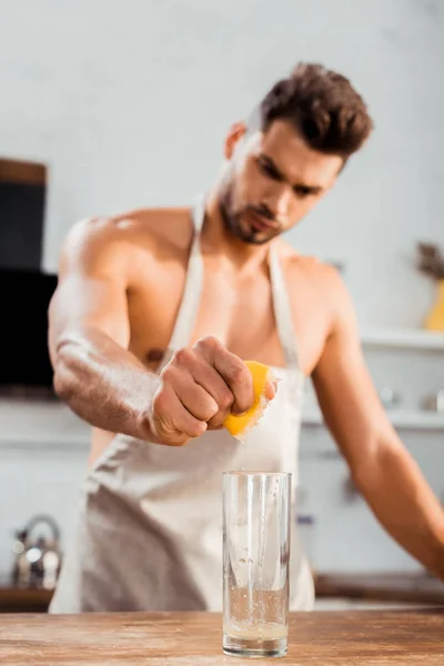 Vue rapprochée du jeune homme à poitrine nue dans un tablier serrant du citron dans du verre — Photo de stock