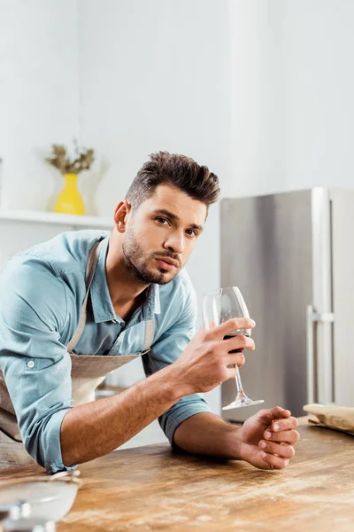 Schöner junger Mann in Schürze hält ein Glas Wein in der Hand und schaut in die Kamera, während er in der Küche kocht — Stockfoto