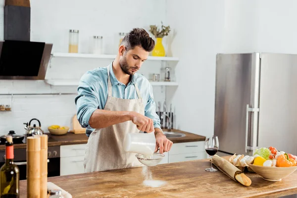 Bel giovanotto in grembiule setacciando farina sul tavolo della cucina — Foto stock