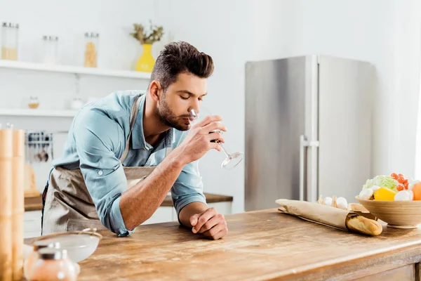 Joven guapo en delantal beber vino mientras se cocina en la cocina — Stock Photo