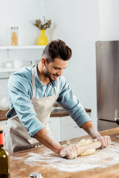 Красивий усміхнений молодий чоловік в фартусі з прокатним штифтом готує тісто на кухні — стокове фото